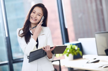 Confident Asian businesswoman in white suit, smiling and talking on mobile phone in a bright office.